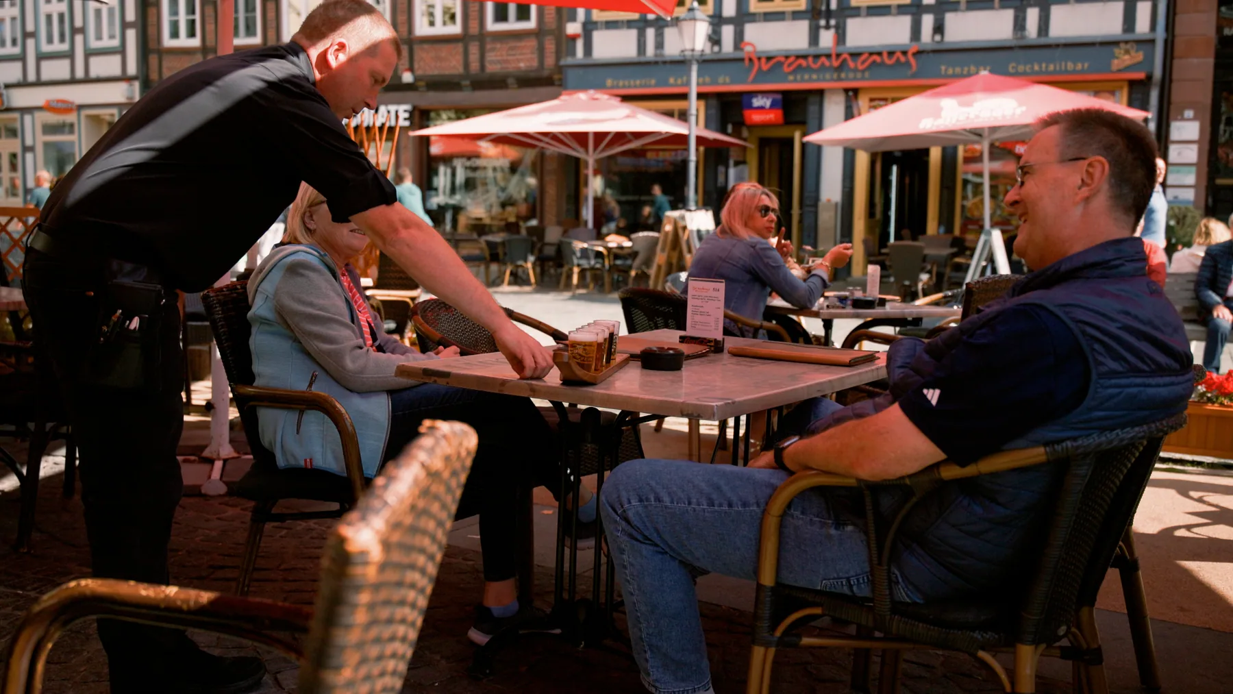 Ein Servicemitarbeiter bedient Gäste mit einem Probierbrett Bier auf der Terrasse unter roten Sonnenschirmen vor dem Brauhaus